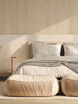 A beige velvet ottoman in front of a bed in a well-lit room, with a matching beige and gray color scheme.