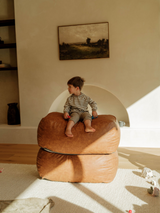 A child sitting on a brown vegan leather ottoman in a room with minimalist decor and a framed picture on the wall.