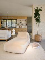 A beige velvet divan with a longer length than usual, placed on a rug in a room with a modern interior design, featuring a kitchen and a large window.