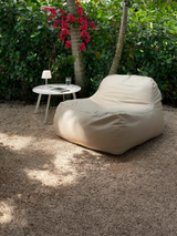 An oat-colored outdoor lounge chair with a flat base and greenery in the background.