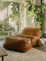 A brown Dume Lounge Chair and Ottoman made of vegan leather, positioned in a room with a large window and plants, with a matching pillow cover and tote bag included.