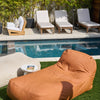 An outdoor divan in a ginger color positioned on grass by a poolside, with lounge chairs and plants in the background.