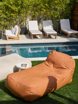 An outdoor divan in a ginger color positioned on grass by a poolside, with lounge chairs and plants in the background.