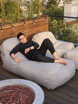 A person relaxing on a gray outdoor divan on a rooftop patio with a fire pit and wooden fence in the background.