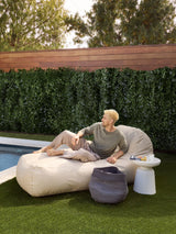 A person relaxing on an beige outdoor divan placed near a poolside with a tote bag beside it.