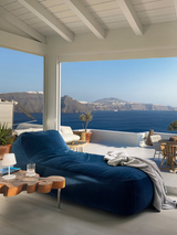 A blue velvet divan lounger with a wooden frame, placed on a balcony with a view of the ocean and mountains in the background. The divan appears plush and comfortable, with a pillow on it, and there are plant pots and a glass of water on the side table.