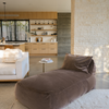 A brown velvet divan lounger placed on a white carpet with a modern living room in the background featuring a low-profile couch and wooden shelving.