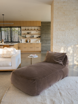 A brown velvet divan lounger placed on a white carpet with a modern living room in the background featuring a low-profile couch and wooden shelving.