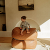 A child sitting on a brown vegan leather ottoman in a room with minimalist decor and a framed picture on the wall.