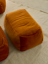 A tangerine-colored velvet ottoman in front of a bed in a stylishly furnished room.