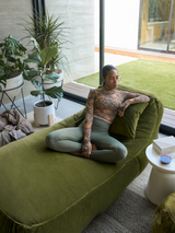 A person relaxing on a green velvet divan in a well-lit room with plants and a window in the background.
